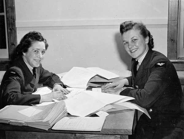 Photographie en noir et blanc – Deux femmes portant un uniforme de l’Aaviation royale du Canada, mais sans casquette, sont assises l’une en face de l’autre à une table couverte de piles de feuilles.